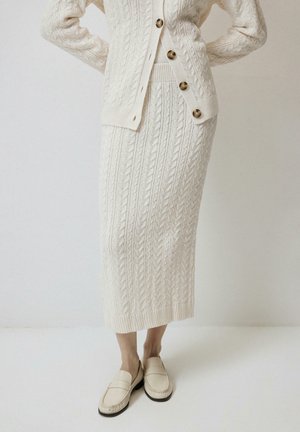 Kremno obarvana pletena krilo iz kabela s pripadajočim jopičem z velikimi rjavimi gumbi in kremnimi loafers na osebi, ki stoji.