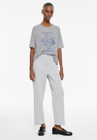 Camiseta de algodón gris con un diseño gráfico azul que dice "Perseverancia y buena suerte", combinada con pantalones de pierna ancha en gris claro y mocasines negros.