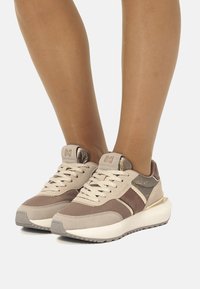 Sneaker beige e marroni in materiali mesh e suede, design con lacci, dettagli metallici e suole in gomma bianca spessa.