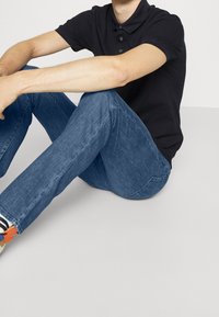 Černé polokošile s límečkem, doplněné modrými džínami. Model sedí na světle šedém povrchu a má na sobě neformální obuv s akcenty.