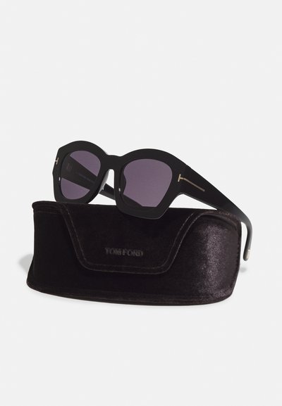Occhiali da sole oversize neri con una montatura spessa, lenti viola e un logo in oro sui lati, appoggiati su un astuccio in velluto nero.