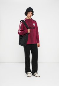 Bordo adīts džemperis ar melniem akcentiem un Adidas logo, apvienots ar melnām platām biksēm, melnu šķērsbiksu somu un baltām kedām.