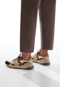 Le sneaker beige e nere presentano un design misto con accenti arancioni, una superficie testurizzata e una suola spessa. Indossate con pantaloni cropped marroni.