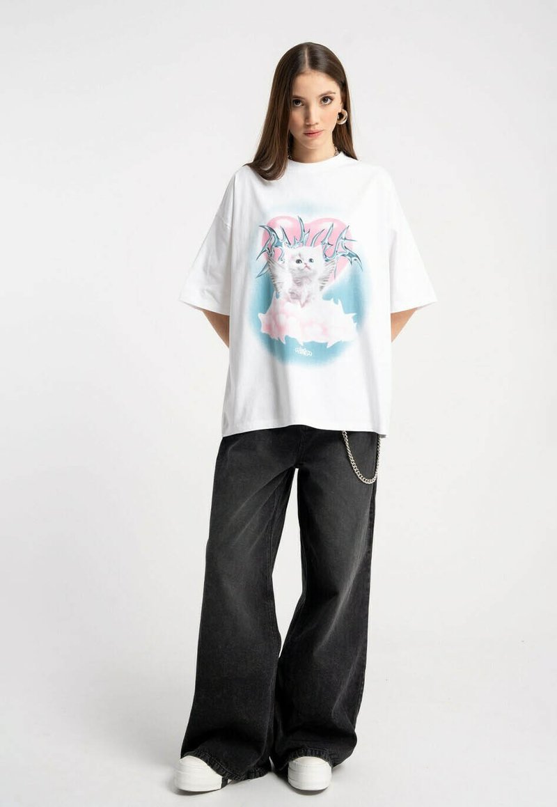 Przesadnie duża, biała koszulka typu T-shirt z grafiką kotka w różowych akcentach, zestawiona z czarnymi spodniami o szerokich nogawkach i białymi trampkami.