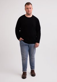 Maglione nero a maniche lunghe, jeans azzurri chiari, scarpe marroni con lacci. Il modello sta in piedi con una mano in tasca, su uno sfondo neutro.