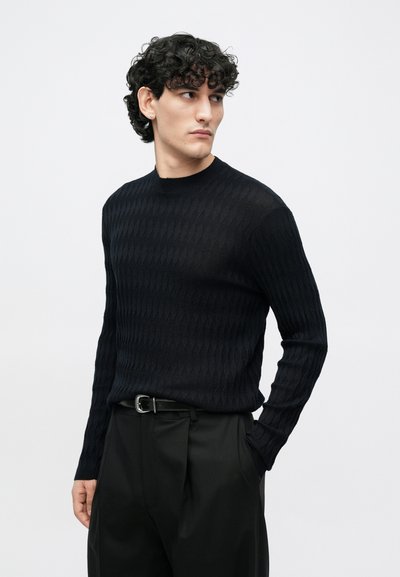 Μαύρο πλεκτό πουλόβερ με υφή, με διακοσμητικό μοτίβο ρόμβου, στρογγυλή λαιμόκοψη και εφαρμοστά μανίκια, συνδυασμένο με μαύρο παντελόνι και ζώνη.