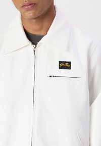 White denim jacket with a pointed collar, front zip closure, and a small black label with yellow text on the chest. Features a zip pocket.