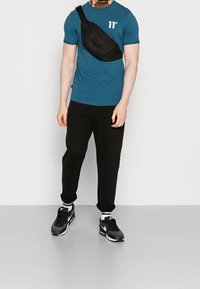Man som bär en teal t-shirt, en svart axelväska, svarta byxor med manschetter, vita strumpor och svarta Nike-sneakers står på ett ljusgrått golv.