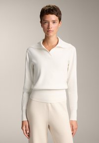 Pulover alb tricotat cu gât larg și tiv cu modele în relief. Mâneci lungi, țesătură texturată și siluetă ajustată. Asortat cu pantaloni bej.