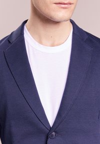 Námořnický modrý blazer s texturovaným povrchem, na jedno knoflíkové zapínání a s klopovými límci, přetažený přes bílý tričko s kulatým výstřihem.