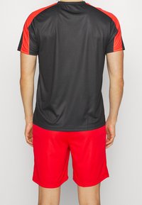 Krátký rukáv, sportovní tričko černé barvy s červenými akcenty na ramenou, v kombinaci s červenými šortkami. Hladký strukturovaný materiál, uvolněný střih.