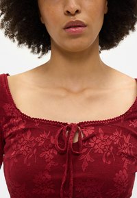 Κόκκινη πλεκτή μπλούζα με υφή λουλουδιών, τετράγωνη λαιμόκοψη και κλείσιμο με κορδόνι μπροστά, με θηλιές στις άκρες.