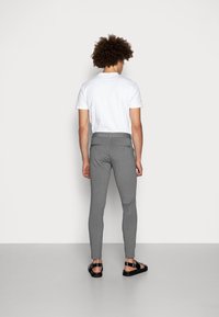 Šedé slim-fit kalhoty s hladkou texturou a páskem, spárované s jednoduchým bílým tričkem a černými sandály. Pohled zezadu.