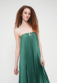 Γυναίκα με μακριά, σγουρά μαλλιά, που φοράει πράσινο φόρεμα χωρίς τιράντες, με πιέτες, μικρή οπή στο ντεκολτέ και χρυσό κουμπί στο λαιμό.