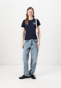 T-shirt di cotone blu marino con un design decorativo, abbinato a jeans chiari con motivi floreali e mocassini neri. Piccola borsa rosa.