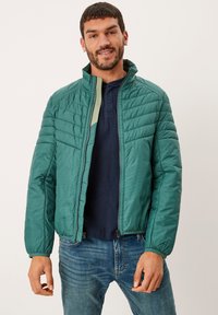 Giacca trapuntata verde con collo alto, zip frontale, polsini elasticizzati e rivestimento interno a costine, indossata sopra una camicia blu navy e jeans.