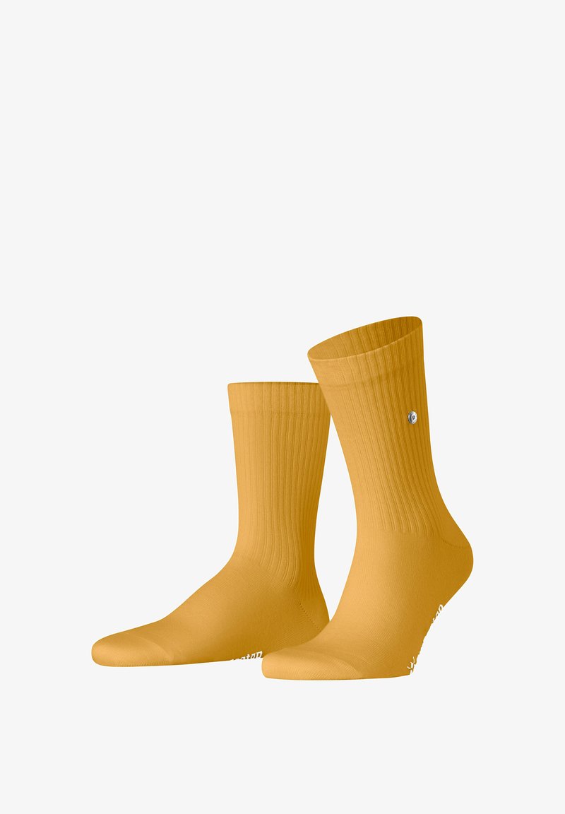 Paire de chaussettes montantes côtelées jaune moutarde avec un petit logo rond argenté sur le dessus et un texte blanc près des orteils.