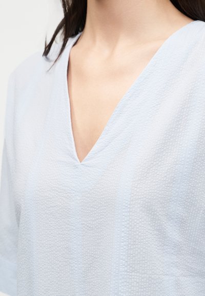 Γυναίκα που φοράει μπλούζα από υφασμάτινη υφή σε ανοιχτό μπλε χρώμα με V-λαιμόκοψη, που αποκαλύπτει τον λαιμό και το πάνω μέρος του στήθους.