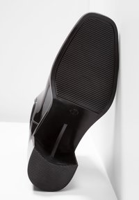 Semelle d'une chaussure à talon haut noire avec une surface antidérapante texturée au niveau du talon et de l’avant-pied, reposant contre une surface blanche.