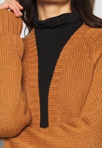 Cardigan de punto en color óxido con un escote en V profundo, puños acanalados y textura gruesa, superpuesto a un cuello de tortuga negro.