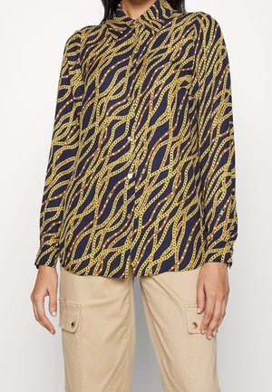 Marineblaue Bluse mit Knopfleiste und einem durchgehenden goldenen Kettenmuster, kombiniert mit beigen Cargohosen mit vorderen Klappentaschen.