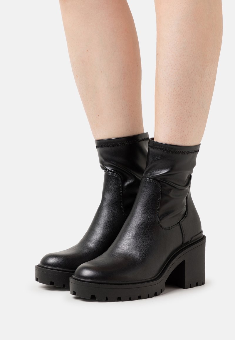 Bottes en cuir noir mi-mollet avec talons carrés épaissis et semelles à crampons portées sur des jambes nues sur un fond clair uni.
