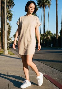 Junge Frau in beigem kurzärmligem Kleid und weißen Turnschuhen, die auf einem sonnenbeschienenen Bürgersteig mit hohen Palmen spaziert.