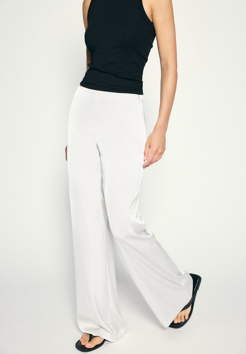 Femme portant un pantalon blanc taille haute à jambes larges, un haut noir sans manches et des tongs noires, debout devant un fond clair uni.