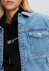 Ljusblå denimjacka med klassisk krage, två framfickor och en sliten textur över en svart huvtröja med vit text.