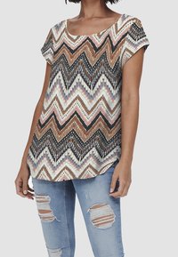 Top a maniche corte con un motivo a zigzag nei colori marrone, nero e pastello. Tessuto morbido, vestibilità rilassata, abbinato a jeans blu strappati.