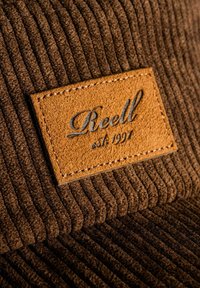 El material de pana marrón presenta una textura de ribetes verticales, con una parche de cuero beige que lleva "Reell est. 1991" en letras estilizadas.