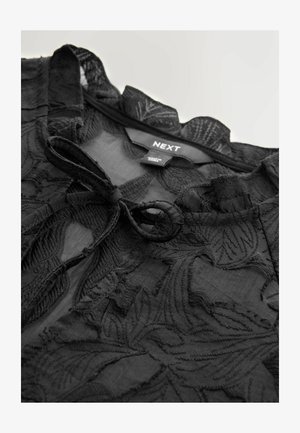 Sort top med blomsterbroderi, let gennemsigtigt stof, frynset halsudskæring og en bindelukning foran. Etiketten er synlig indeni.