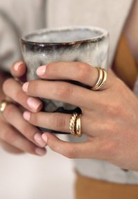 Tasse en céramique avec une finition texturée noir et blanc tenue par des mains portant trois anneaux en or. Surface lisse et vernis à ongles rose doux.