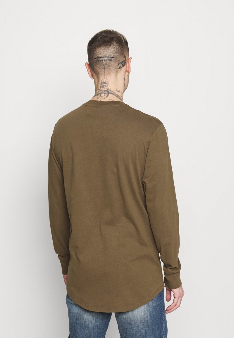 Homme aux cheveux courts avec plusieurs tatouages au cou, portant une chemise olive à manches longues et un jean bleu, tourné de dos sur un fond uni.