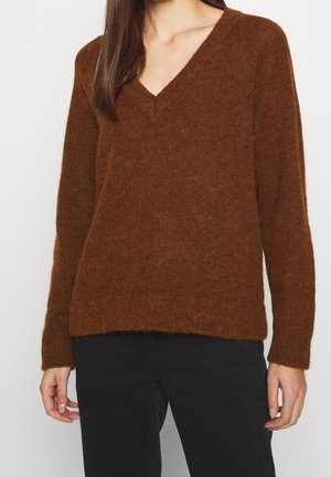 Femme portant un pull marron à col en V en tissu duveteux à manches longues et un pantalon noir, vue des épaules jusqu’aux hanches sur un fond uni.