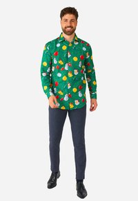 Camicia verde a maniche lunghe con una stampa natalizia di pupazzi di neve, Babbo Natale e ornamenti su sfondo di pini; abbinata a pantaloni navy testurizzati e scarpe nere.