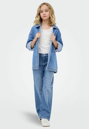 Kind mit blondem Haar, das ein weißes Oberteil, eine übergroße Jeansjacke, Jeans mit weitem Bein und weiße Turnschuhe trägt, steht vor einem einfarbigen Hintergrund.