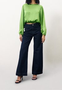Blouse longue à manches longues en satin vert avec ourlet froncé, associée à un jean large taille haute en denim foncé et des sandales à talons noires.