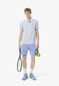 Ljusblå polo med rutmönster, vit krage, i kombination med lavendelfärgade shorts. Håller en grön tennisracket och en påse med tennisbollar.