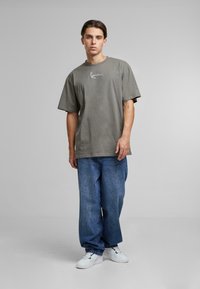 T-shirt en coton gris surdimensionné avec un logo blanc, associé à un jean en denim bleu ample et des sneakers blanches, debout sur un fond gris.