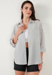 White and navy striped button-up shirt with a collar, roll-up sleeves, and a front pocket. Worn over a white crop top and black shorts.