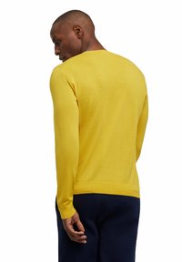 Gelbes langärmliges Sweatshirt mit rundem Ausschnitt, gerippten Bündchen und Saum; glatte Textur und enganliegendes Design. Über marineblauen Hosen getragen.