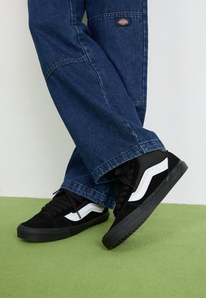 Schwarz-weiße Sneaker mit einer strukturierten Oberfläche, die ein markantes weißes Wellenmuster aufweisen, kombiniert mit lockeren blauen Jeans.