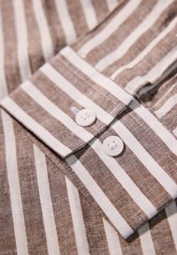Tela a rayas marrón y blanca con un cuello puntiagudo, que presenta dos botones blancos en la abertura. La textura parece ser tejida y ligeramente áspera.