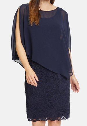 Kobieta ubrana w granatową koronkową sukienkę z przezroczystą asymetryczną nakładką i bezrękawnym krojem, stojąca na jednolitym tle.