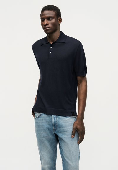 Homme portant un polo noir à manches courtes avec trois boutons et un jean bleu clair, debout devant un fond blanc uni.