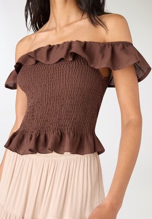 Donna che indossa un top marrone a spalle scoperte con arricciature e bordi arruffati abbinato a una gonna plissettata beige chiaro, mostrata dalle spalle alla vita.