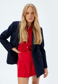 Blazer navy de dos botones sobre una camisa de punto rojo y una falda roja brillante. Textura suave, ajuste a medida y contraste de color vibrante.