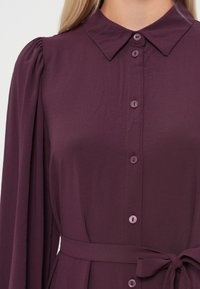Rochie mov cu guler, cu mâneci pliate și curea în talie. Material moale, cu o textură fină și un luciu subtil.