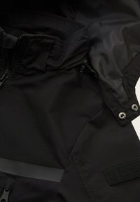 Schwarze Outdoor-Jacke mit glatter Textur, Reißverschlusstaschen, verstellbaren Bündchen, reflektierendem Streifen und Markenpatch-Detail.